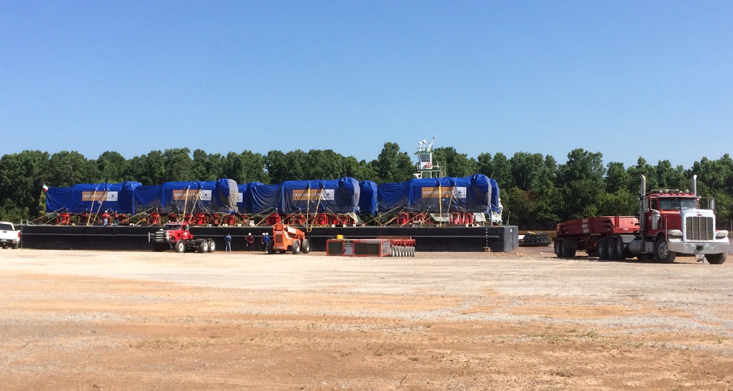 Large industrial equipment covered with blue tarps on an elongated trailer, parked in a dirt lot with a red tractor truck attached.