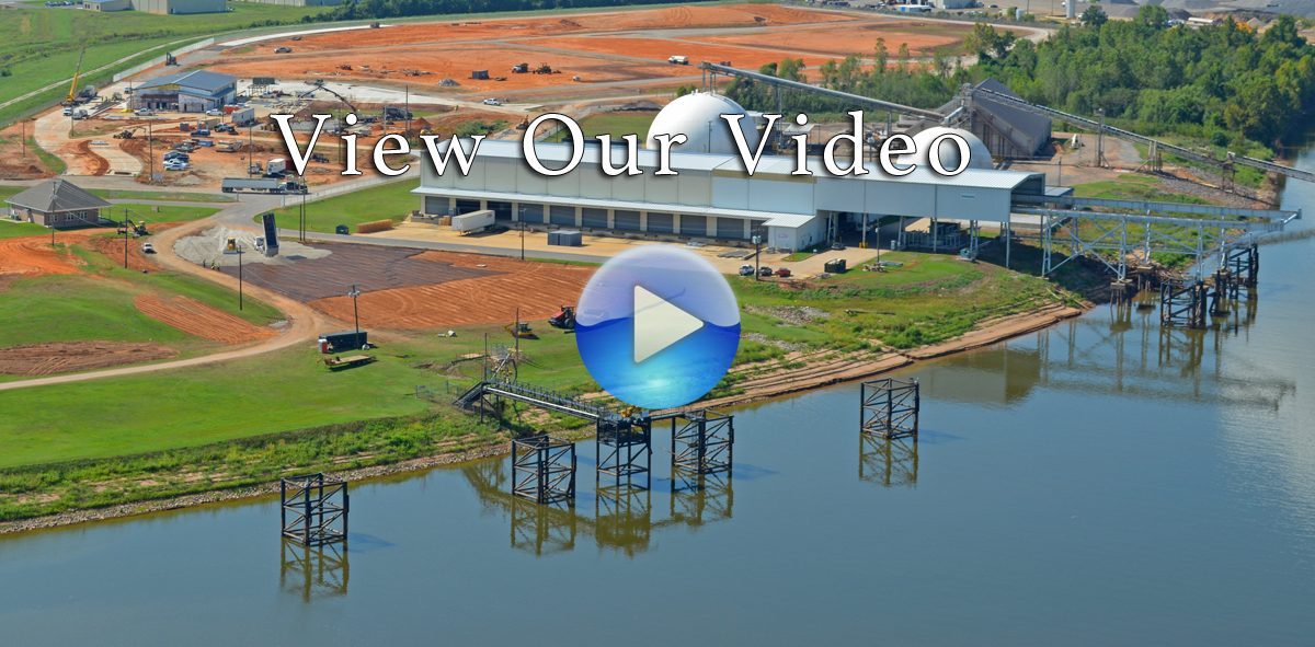 Aerial view of an industrial facility near a body of water with a play button overlay inviting viewers to watch a video.
